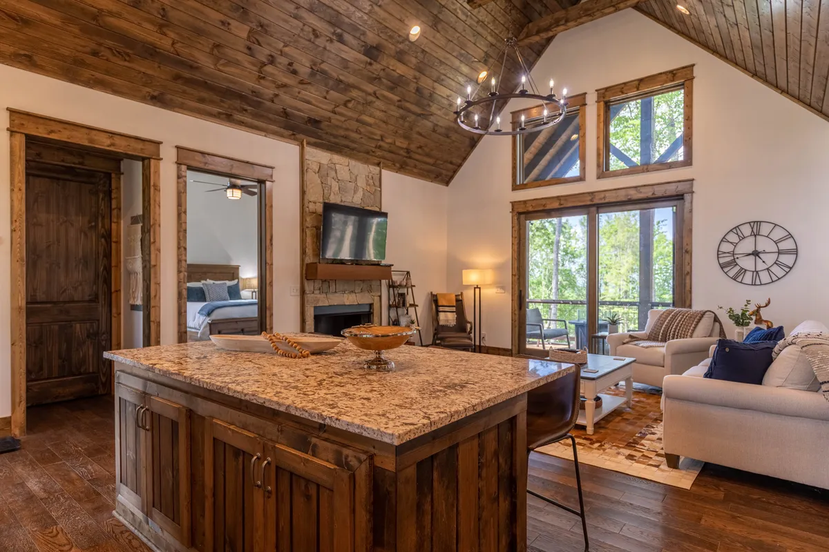 A spacious open-layout featuring a granite kitchen island in the foreground, a high ceiling with rustic wooden beams, expansive windows letting in natural light, and a comfortable seating area that blends kitchen, dining, and living spaces. Nature views are visible through the windows, reinforcing the mountain setting.