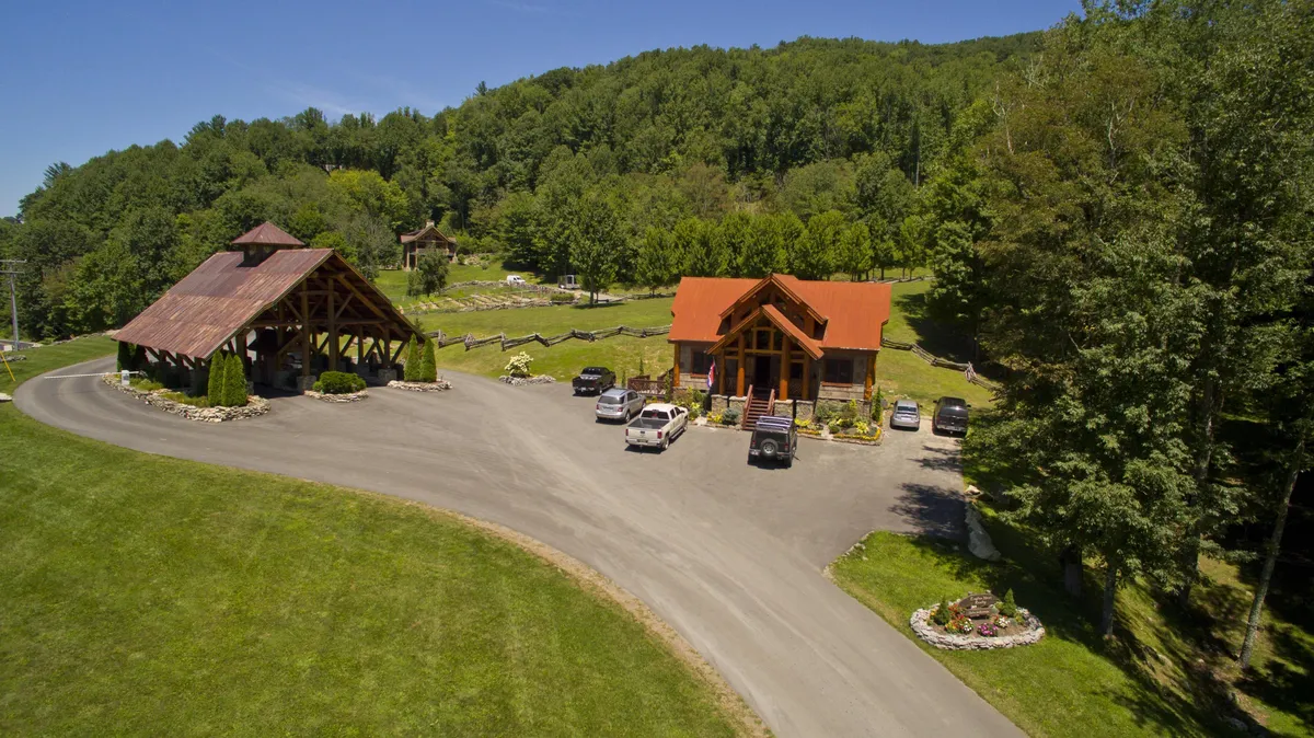 This exterior shot captures log cabin-style buildings surrounded by lush greenery, with a clear view of a circular driveway and ample parking area. The scene radiates warmth and welcome, suggesting an inviting arrival experience for guests.
