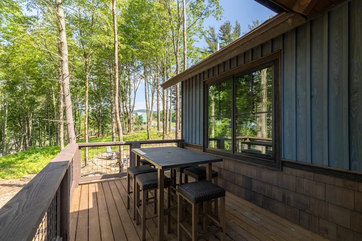 A modern wooden deck with sleek furniture, backed by tall trees and a large window that connects the interior to the exterior. The arrangement is perfect for sunset dinners or peaceful mornings, with a sense of openness to the landscape and a seamless indoor-outdoor transition.