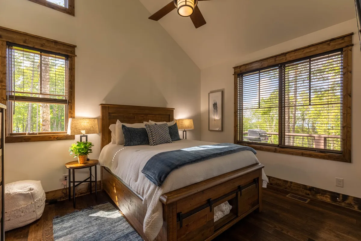 A serene bedroom with a wooden bed frame, large windows overlooking the forest, decorative pillows, a bedside table lamp, and a green plant. Natural light fills the space, creating a tranquil atmosphere perfect for unwinding.