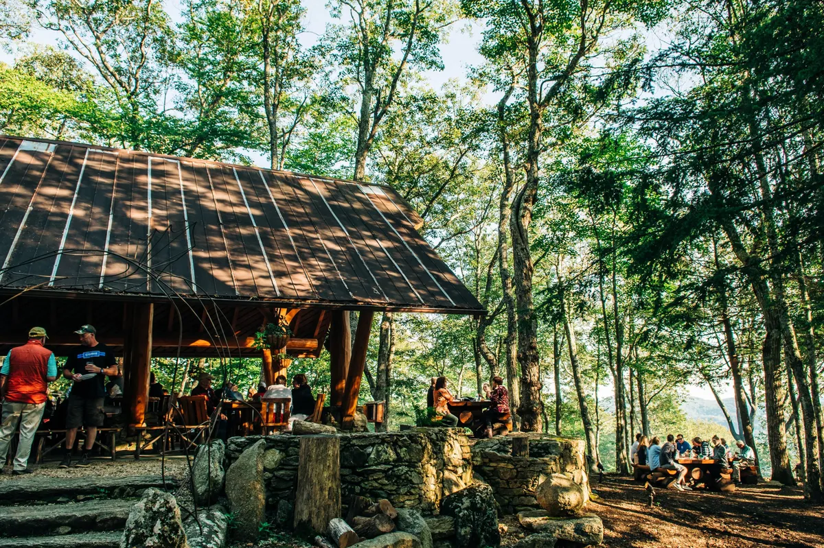 A rustic wooden structure surrounded by a lush green canopy, with a lively group of people gathered in a natural stone seating area. The relaxed, social atmosphere hints at the communal energy of a winery patio or tasting room experience.