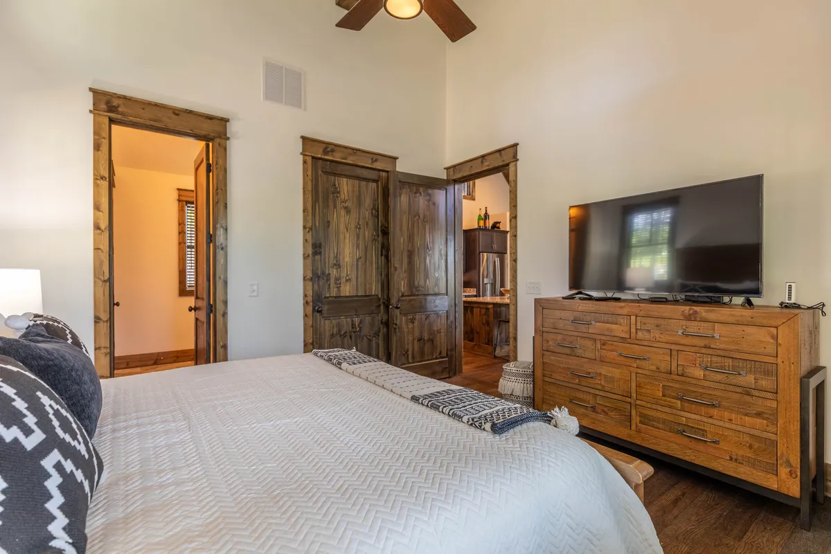 A cozy bedroom featuring a bed with patterned bedding, wooden furniture, and a TV mounted on the wall. Open doorways connect the room to the rest of the suite, creating a spacious yet intimate environment.