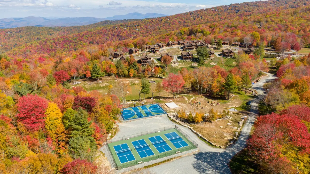 A vibrant exterior view showcasing colorful autumn foliage, rustic cabins, and tennis courts nestled in the mountains. The surrounding landscape pops with the hues of fall, highlighting the natural beauty and variety of outdoor activities available.