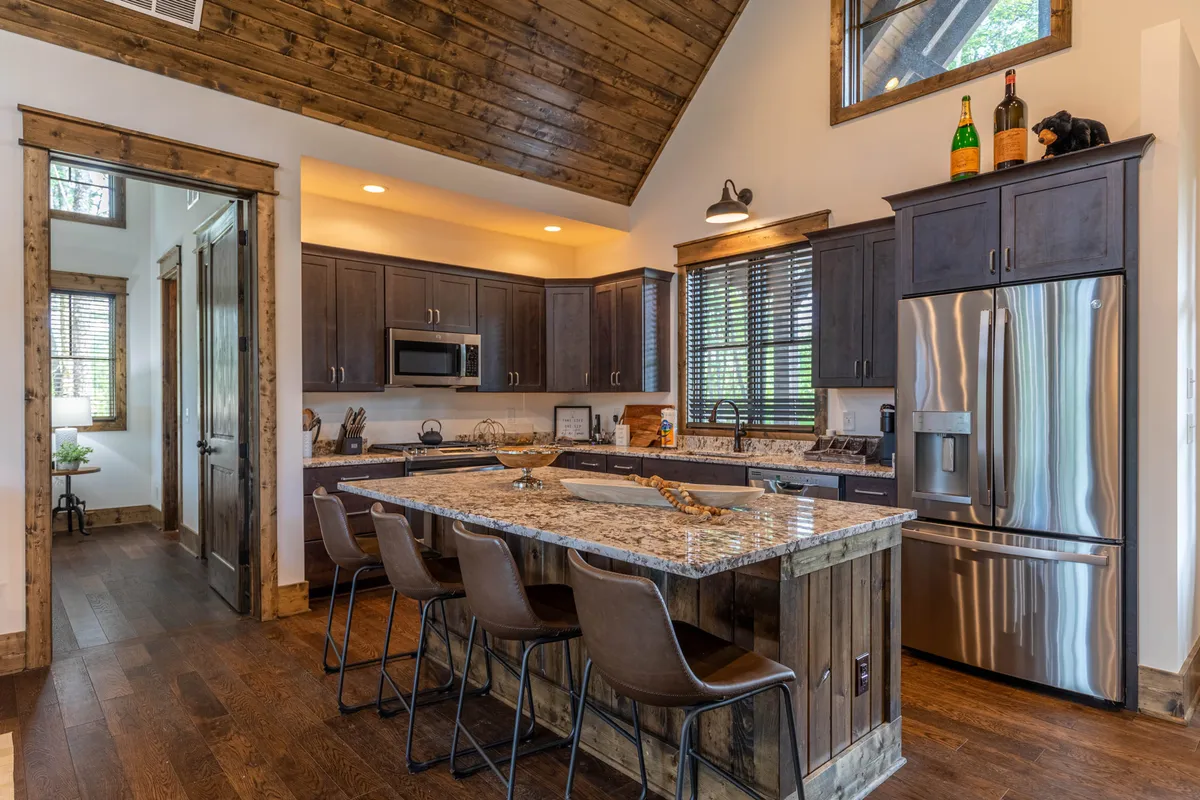 A beautifully designed kitchen featuring a gleaming granite countertop, warm wooden cabinets, an inviting island with seating, large windows bringing in natural light, and decorative bottles adding a homely touch. The open layout visually connects the kitchen with the dining and living areas.