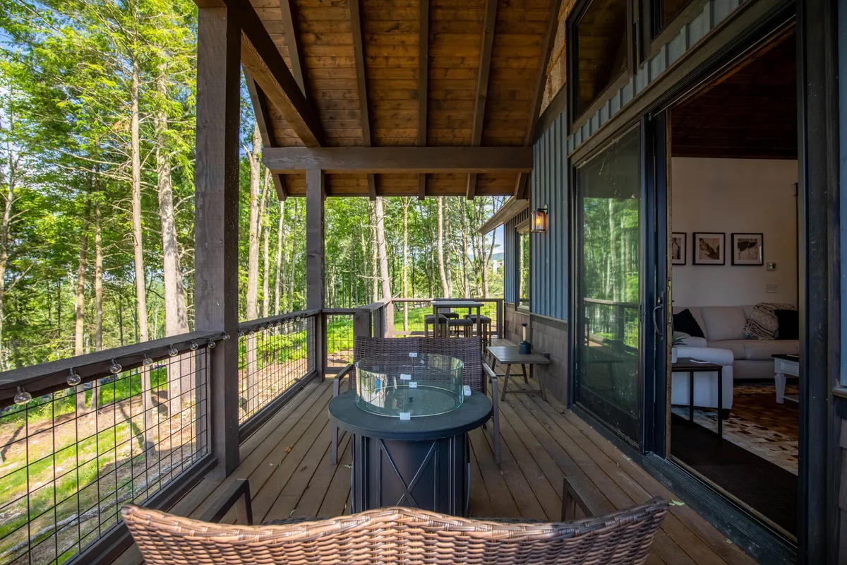 This image presents a wooden deck with comfortable outdoor seating, positioned to overlook the surrounding treetops. Through the open glass doors, you can glimpse the inviting indoor living space, visually connecting the inside and outside. The setting is tranquil and clearly designed for relaxation, and the angle draws attention to how the deck serves as a natural extension of the home's living area.