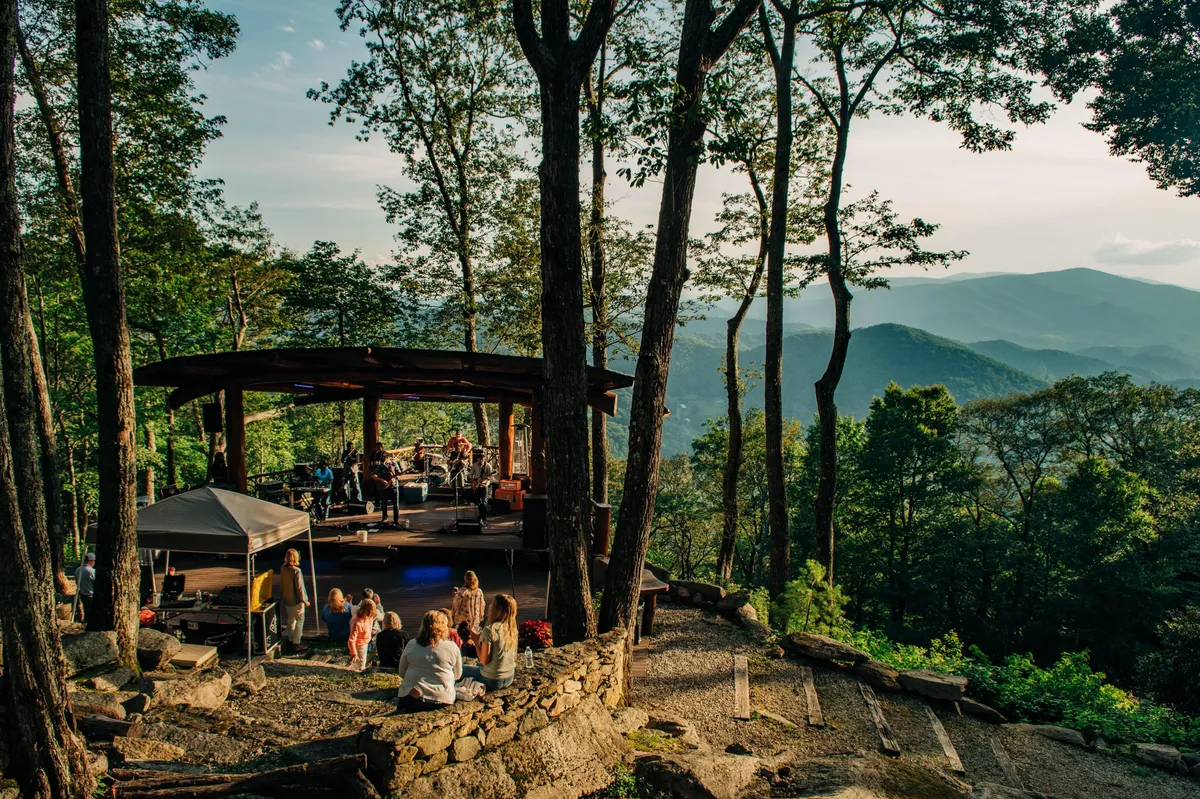 The photo features an outdoor stage set against the scenic backdrop of distant mountains, with people gathering around and trees framing the space. The natural beauty, crowd, and stage evoke the essence of communal outdoor concerts and mountain experiences.