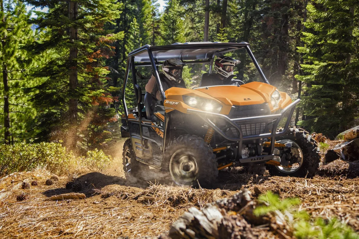 Two riders on an ATV navigate a scenic trail surrounded by dense trees. The scene is lively and full of energy, showcasing off-roading as an adventurous way to explore the mountainous landscape.
