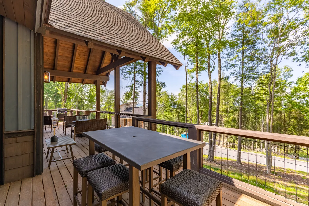 A spacious wooden deck surrounded by trees, featuring an outdoor dining table beneath a shaded roof. The arrangement suggests both shade and openness, making it ideal for dining, socializing, and relaxing while immersed in nature. The inviting setup encourages guests to enjoy meals or drinks with mountain air and tranquil surroundings.