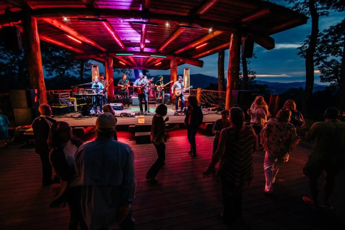 This image captures a lively outdoor concert scene with musicians performing on stage, a crowd dancing, and vibrant, colorful lighting illuminating the event. The atmosphere is energetic and celebratory, perfectly reflecting the communal spirit of live music events at Great Camp.